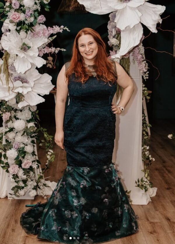 A woman with long red hair, in the striking Rittenhouse Dress Preorders featuring a dark green floral design, smiles in front of an indoor floral archway adorned with large white flowers and greenery on wooden flooring.